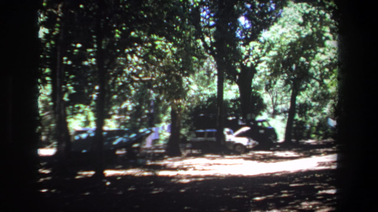 1969 Pretoria home-movie frame showing cars in a wooded lot, including a white car with its hood open beside a dark van-like vehicle.