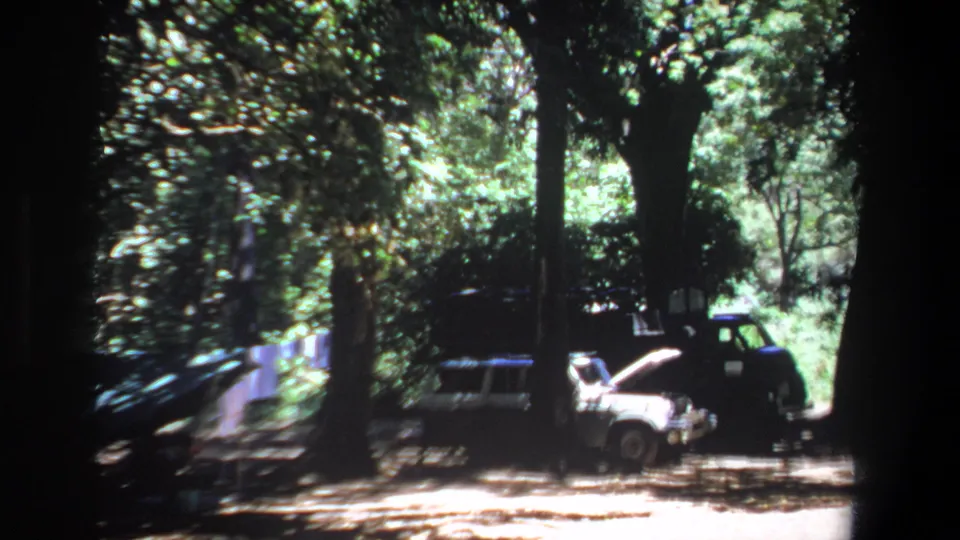 1969 Pretoria home-movie frame with a closer view of parked vehicles under trees, centered on a white car with its hood open.