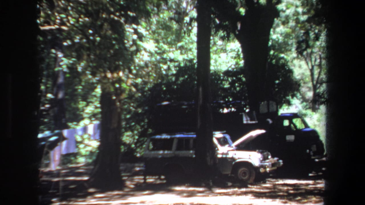 1969 Pretoria home-movie frame showing a light station wagon with its hood open near a dark truck under tall trees.