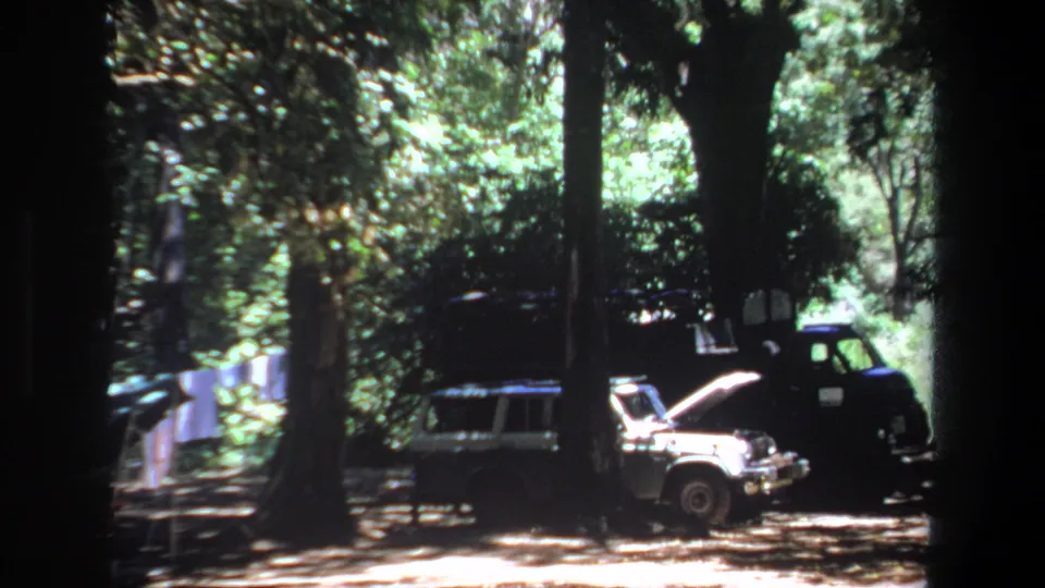 1969 Pretoria home-movie frame showing a light station wagon with its hood open near a dark truck under tall trees.