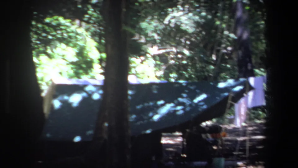 The final frame holds on the campsite setup, with a dark tarp or tent, laundry line, trees, and bright foliage in the background.