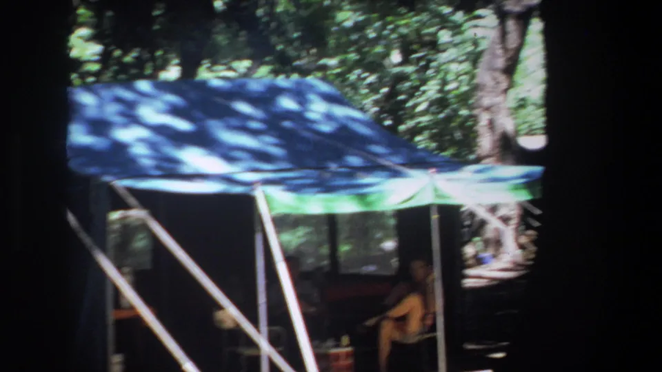 Pretoria 1969 home-movie frame showing people seated under a blue and green canopy tent in a wooded camping area.
