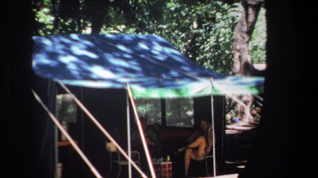 1969 Pretoria home-movie frame showing a blue and green canopy tent in woodland shade with a seated camper inside.