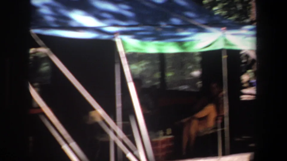 1969 Pretoria 8mm frame showing a canopy shelter, angled support poles, and a camper seated in the shaded interior.