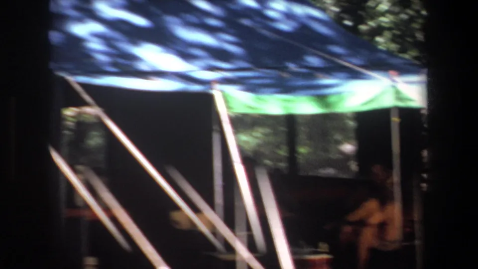 1969 Pretoria camping frame with a blue and green canopy tent, tree shade, support poles, and a seated camper at right.