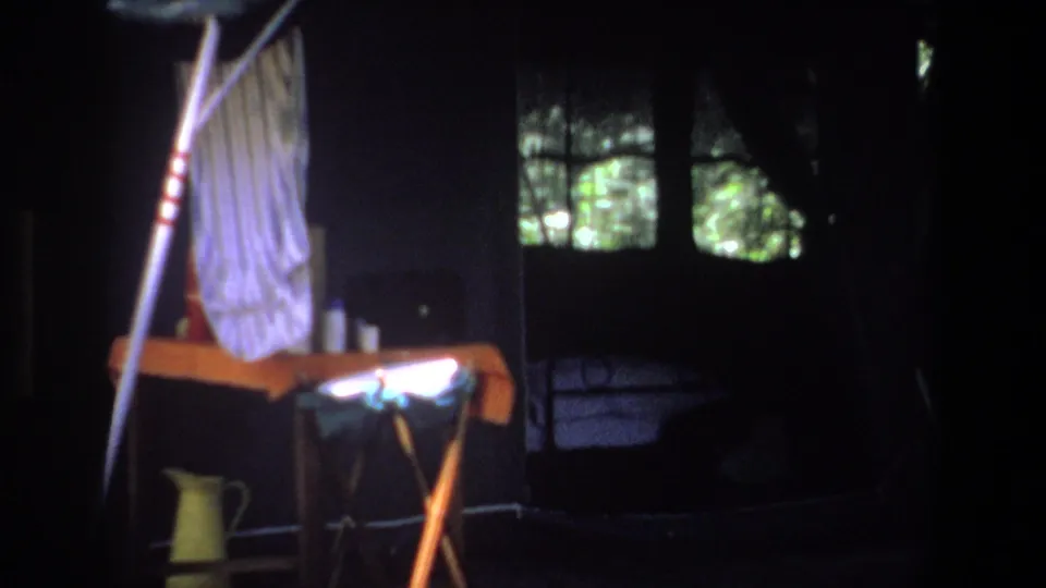 Pretoria 1969 camp interior with a wooden table, yellow jug, hanging cloth, and dark mesh panels facing the trees.