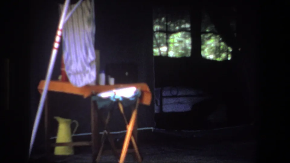 Late-1960s 8mm frame of the Pretoria campsite interior, showing a table, yellow jug, tent pole, and green foliage through dark openings.