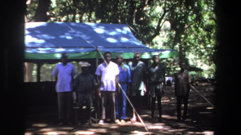 A small group of men stands close together near the blue and green canopy in wooded 1969 Pretoria home-movie footage.