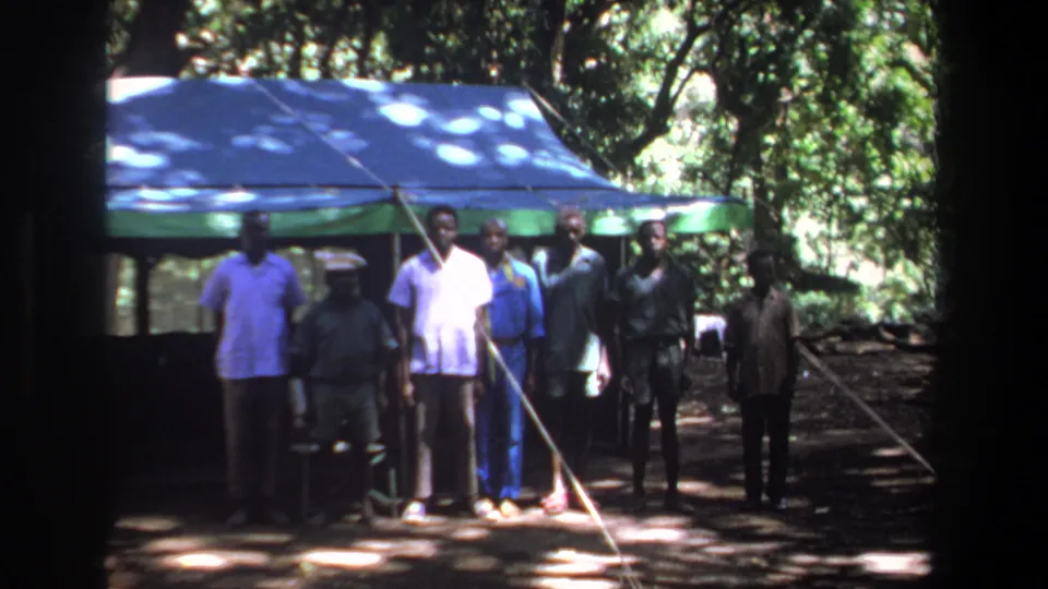 Seven men face the camera in front of a blue and green canopy, surrounded by trees in 1969 Pretoria footage.