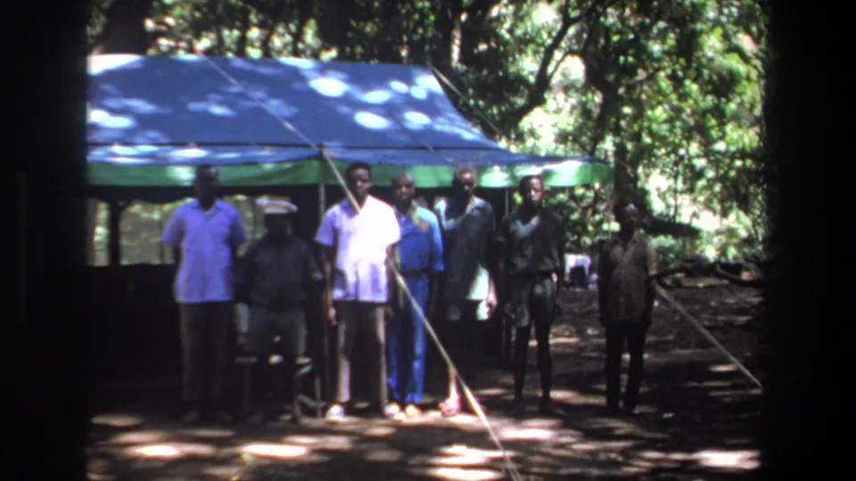 Men stand in a loose line under a blue canopy in 1969 Pretoria footage, with wooded shade behind them.