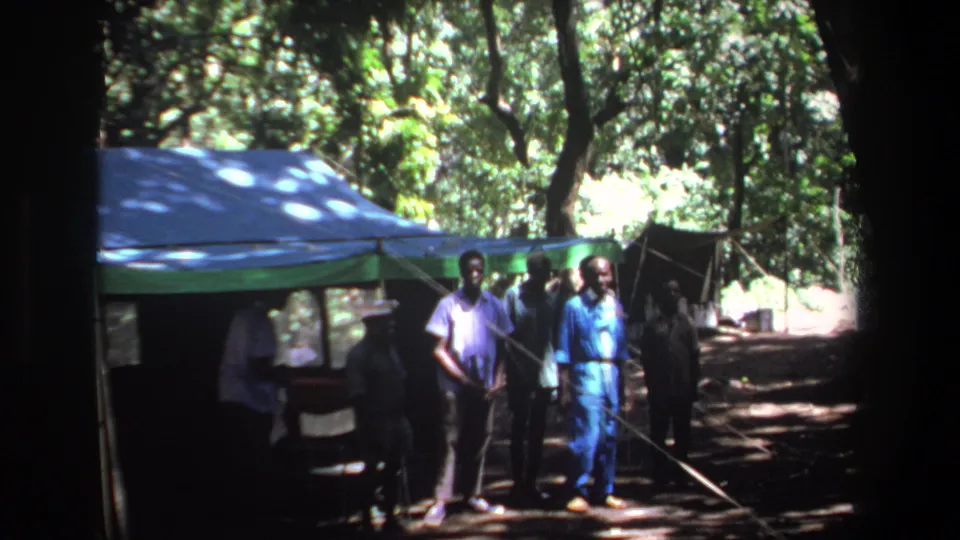 1969 Pretoria frame with men standing near a blue tent canopy in a wooded, sun-dappled outdoor area.