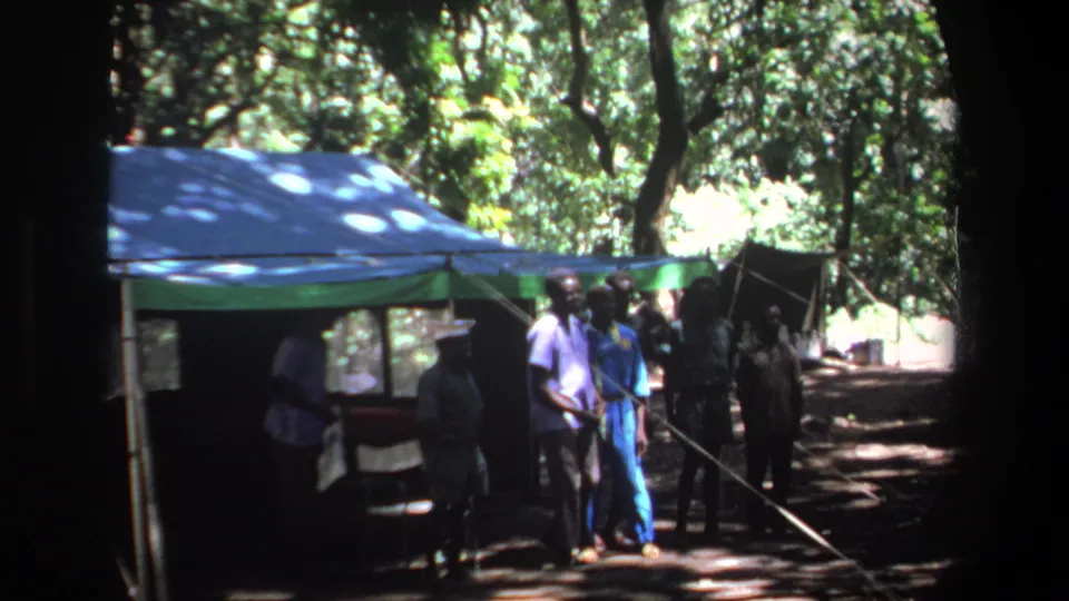 1969 Pretoria home-movie frame showing men around a tent in deep shade with bright trees beyond.
