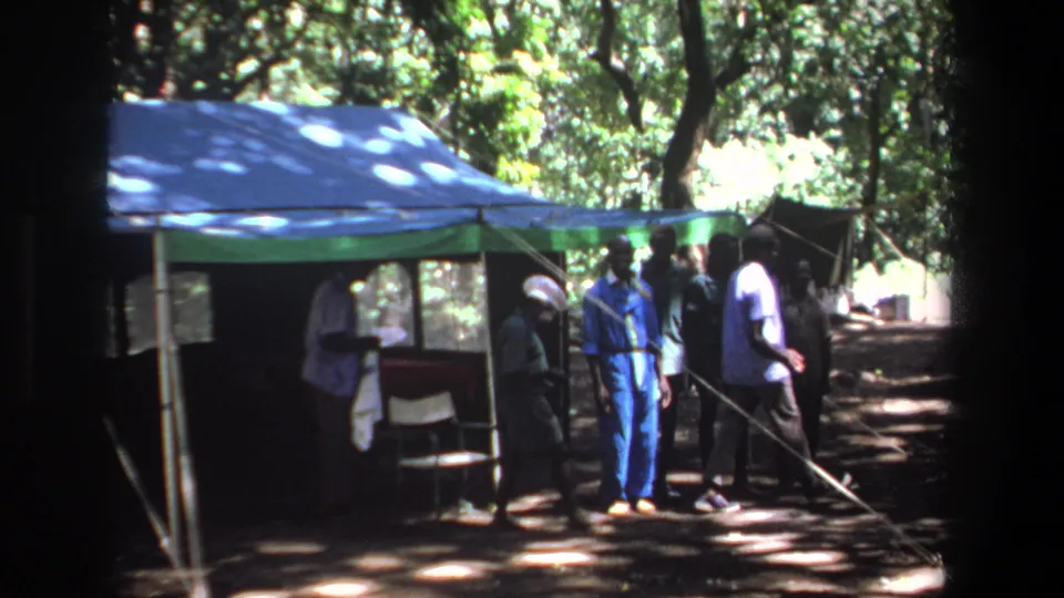1969 Pretoria frame showing men moving around a blue and green tent in a tree-covered clearing.