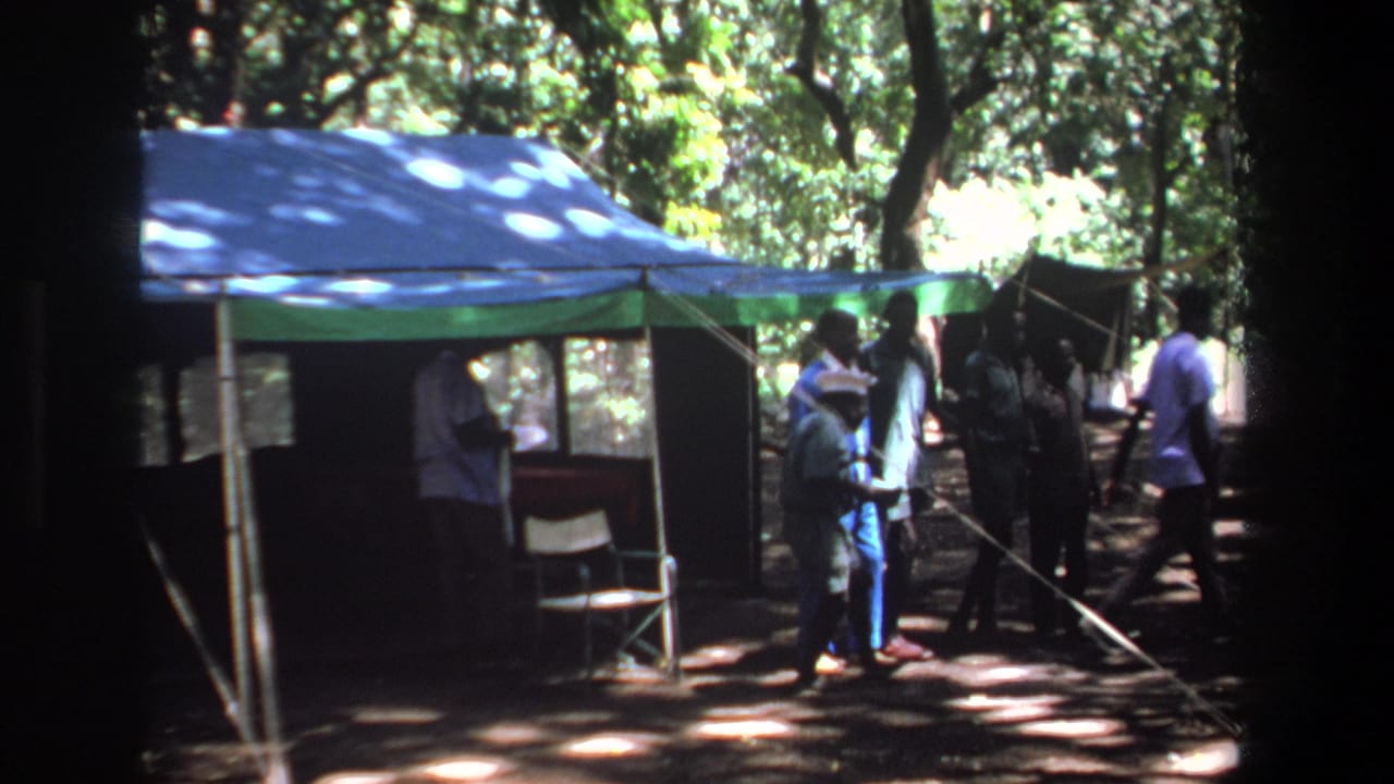 1969 Pretoria home-movie frame showing men gathered by a tarp-covered camp shelter under trees.