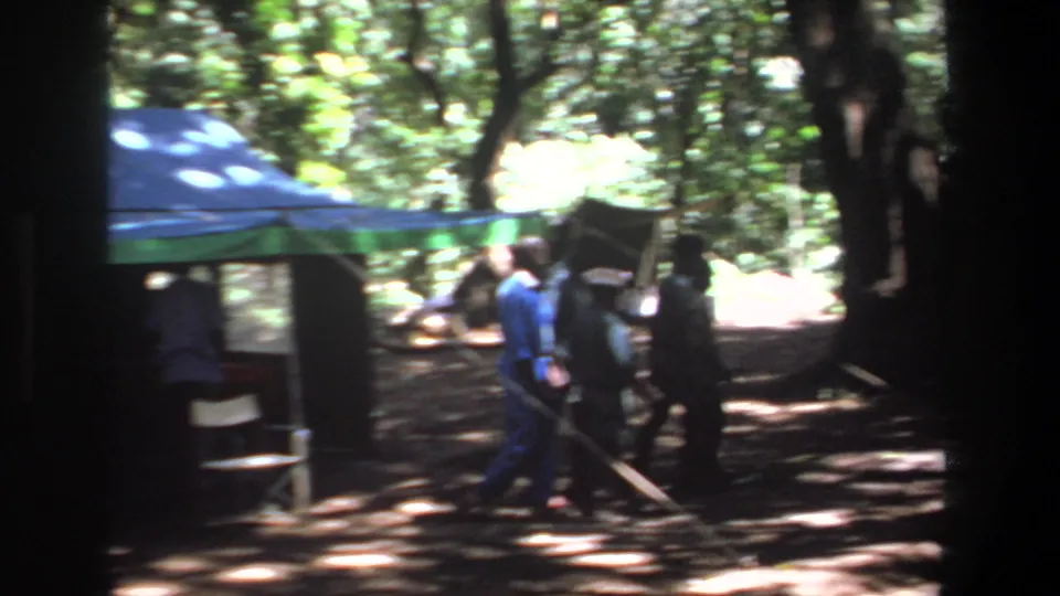 1969 Pretoria frame with men walking through a wooded campsite beside a blue and green tarp shelter.