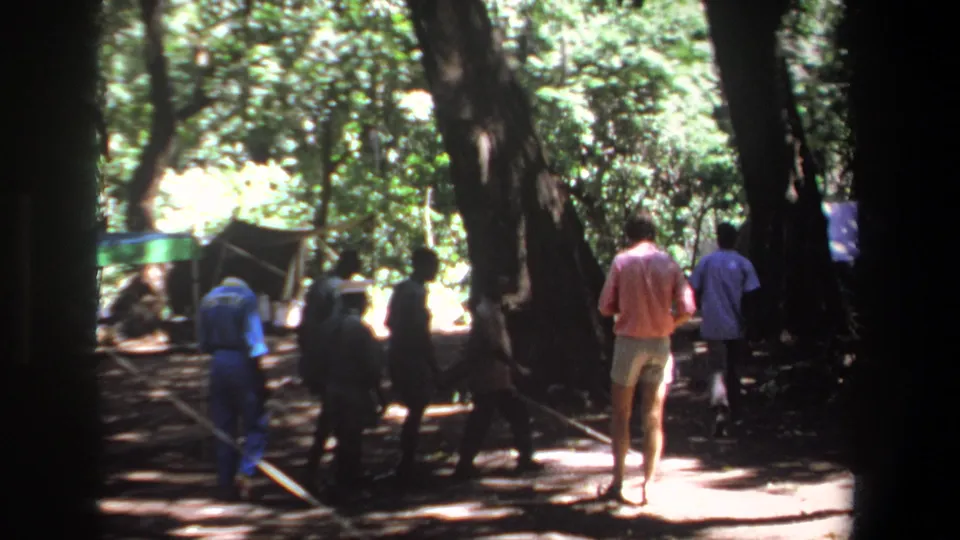 1969 Pretoria home-movie frame showing men walking away through a shaded forest campsite.