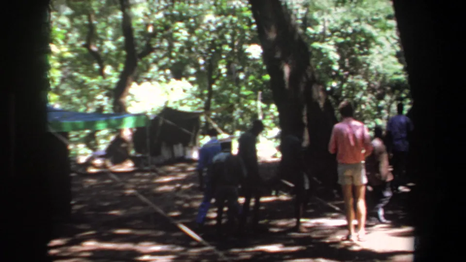 1969 Pretoria frame showing men walking through a forest campsite with tarp shelter visible in the background.