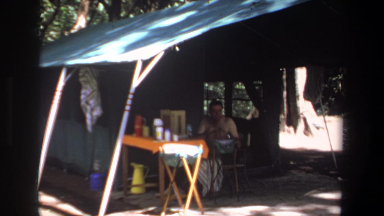 Man seated under a blue tarp beside a folding table with containers and a yellow pitcher.