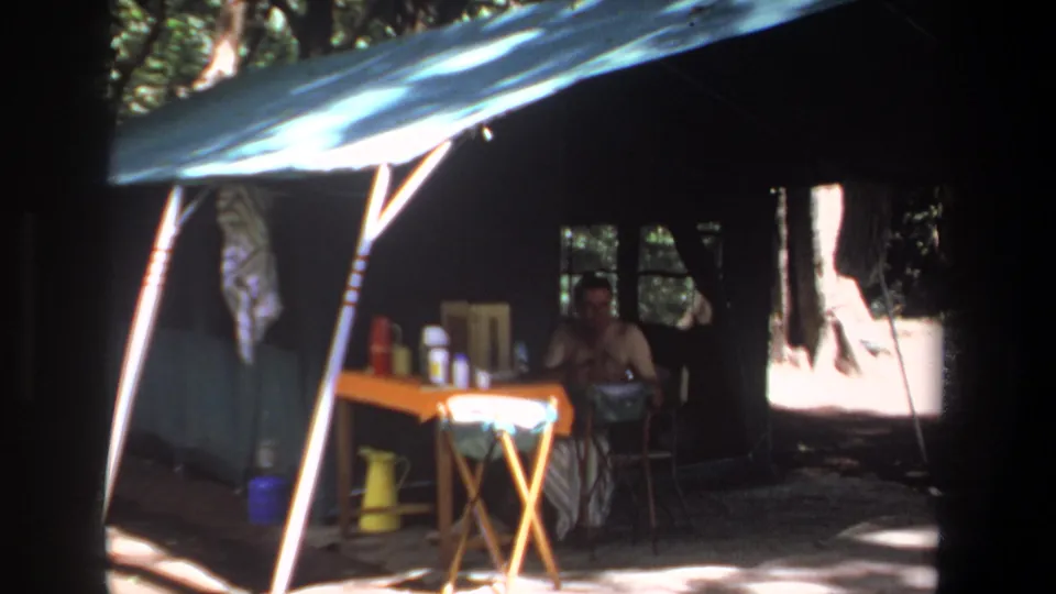 In the Pretoria 1969 campsite, a man sits in shade beside a small table arranged with containers and a yellow pitcher.