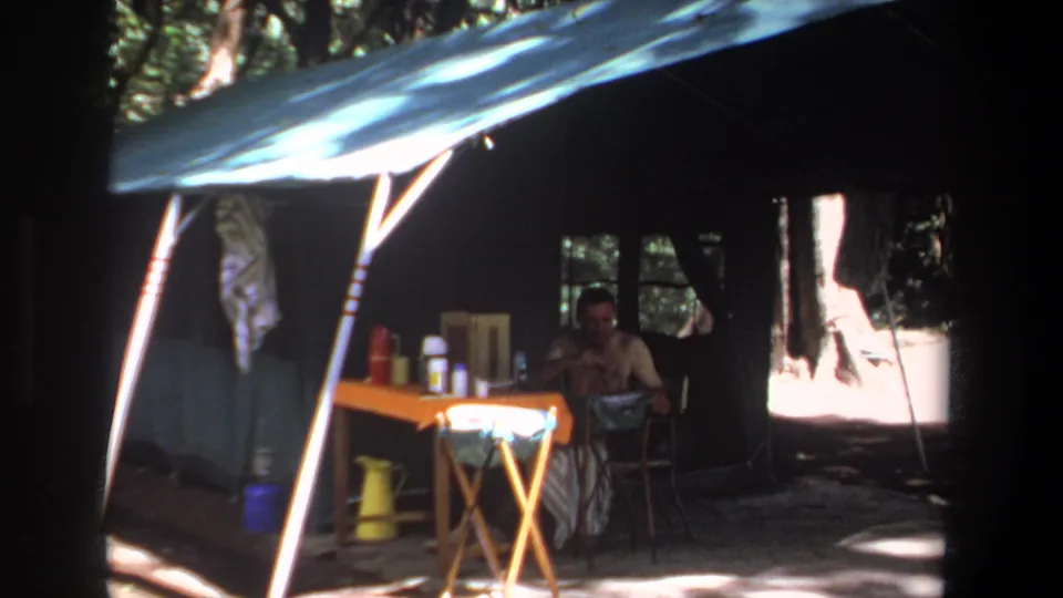 The 1969 Pretoria home movie shows a camper seated in deep shade near a folding table beneath a blue tarp shelter.