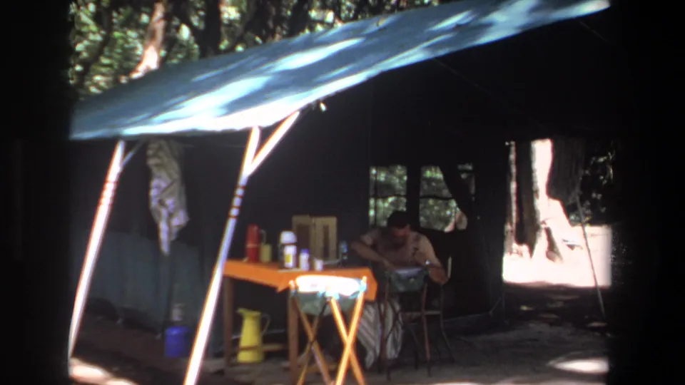 A camper in the Pretoria 1969 campsite bends toward the table under a blue tarp, with containers and a yellow pitcher nearby.