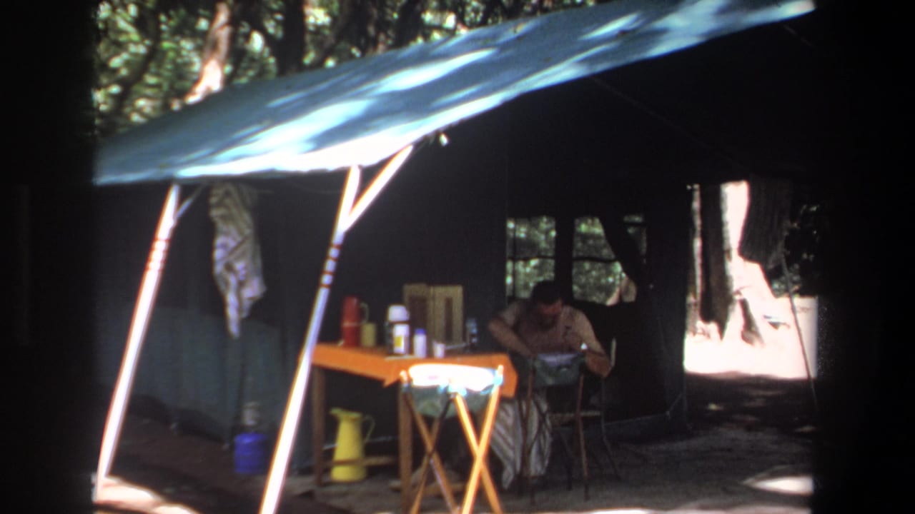1969 Pretoria home-movie frame showing an adult man inside a shaded canvas tent with camping items on a small orange table.