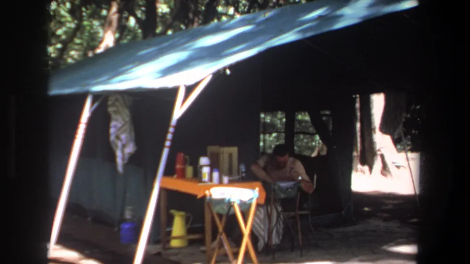 1969 Pretoria home-movie frame showing an adult man inside a shaded canvas tent with camping items on a small orange table.