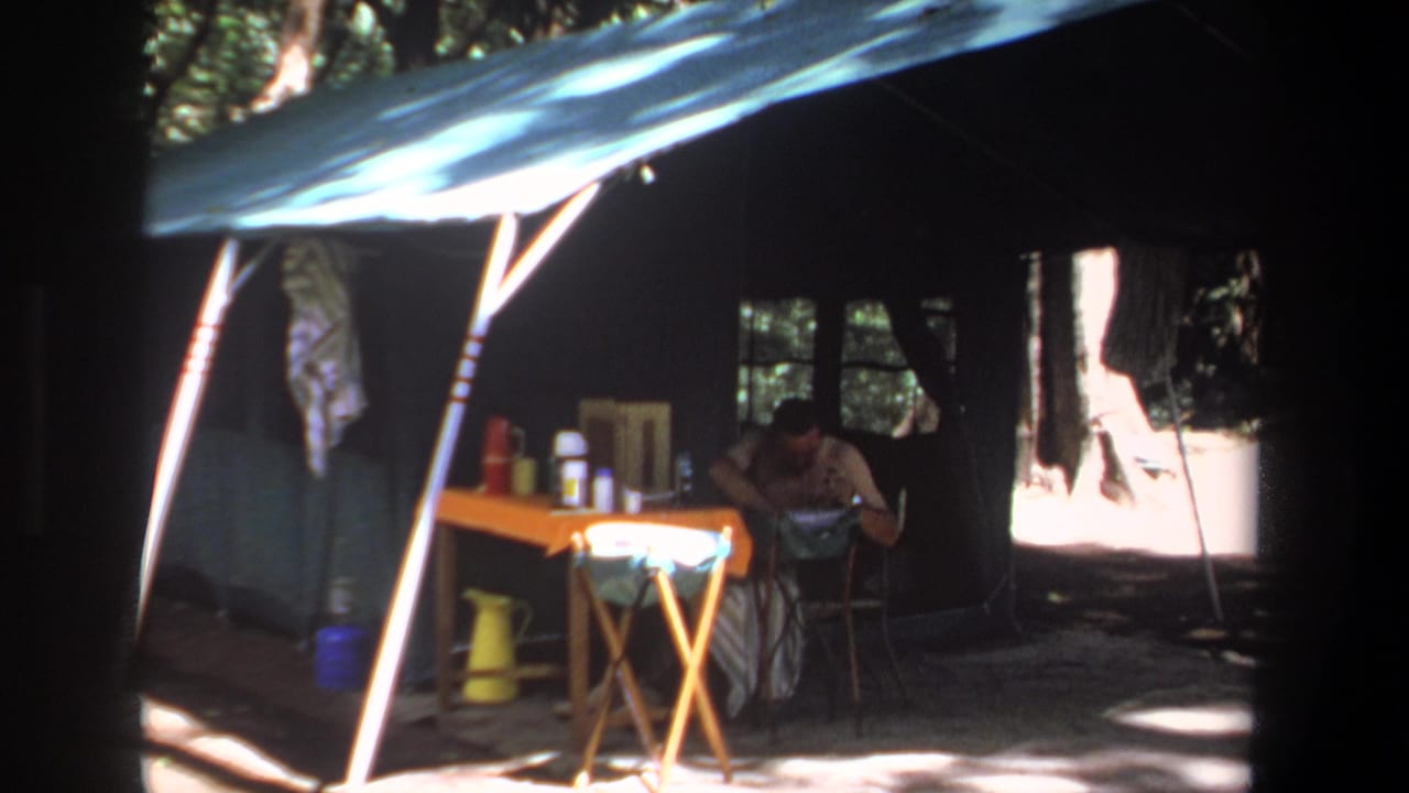 Adult man seated at a small table inside a canvas tent in wooded shade.