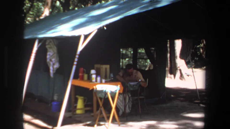 1969 Pretoria 8mm frame showing a man leaning at a folding table inside a canvas tent surrounded by camping supplies.