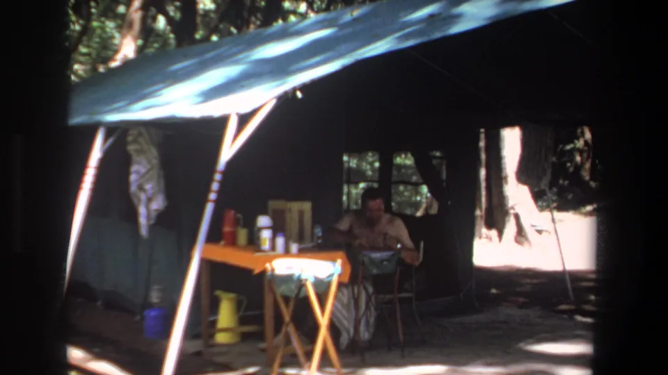 1969 Pretoria home-movie still of an adult man in a shaded canvas tent beside an orange table, thermoses, cups, and a yellow pitcher.