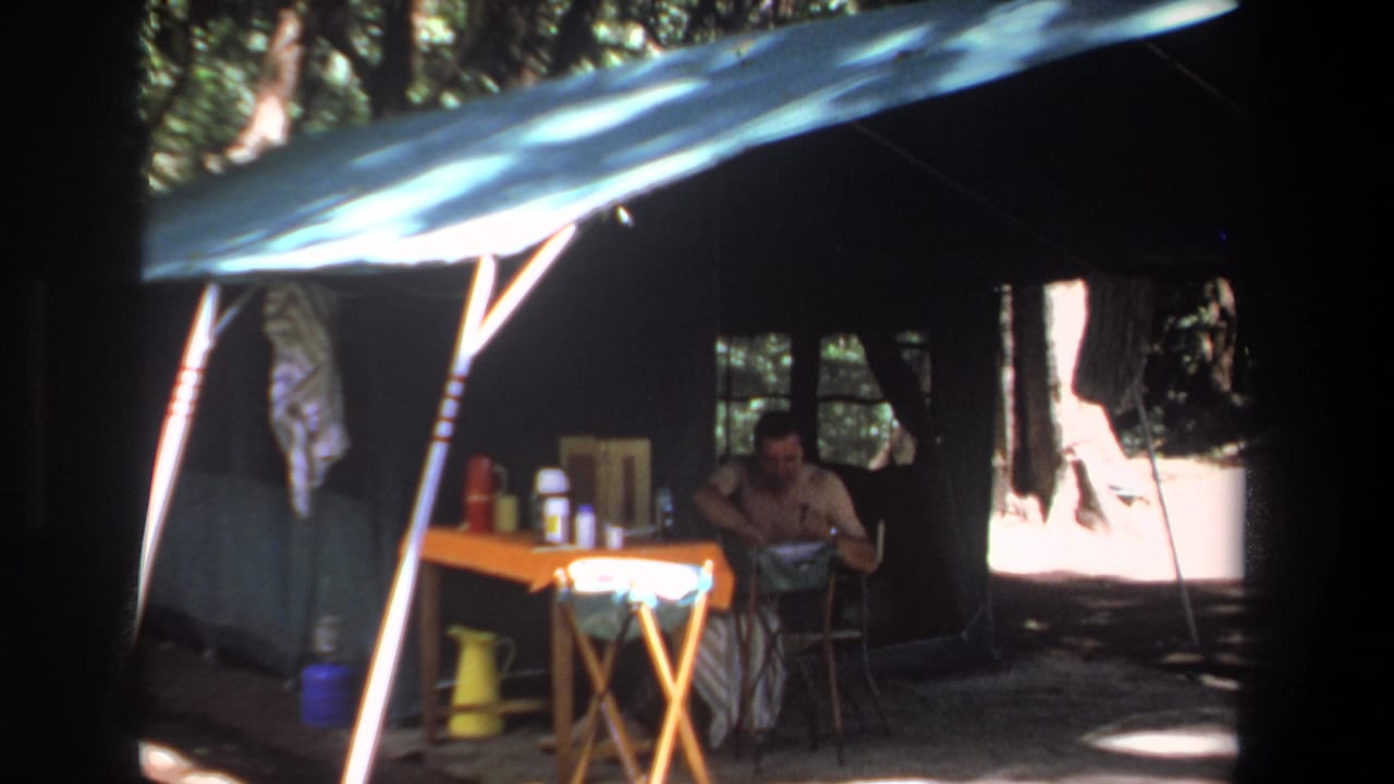 In 1969 Pretoria home-movie footage, a man sits inside a dark forest tent near a table holding thermoses and a yellow pitcher.