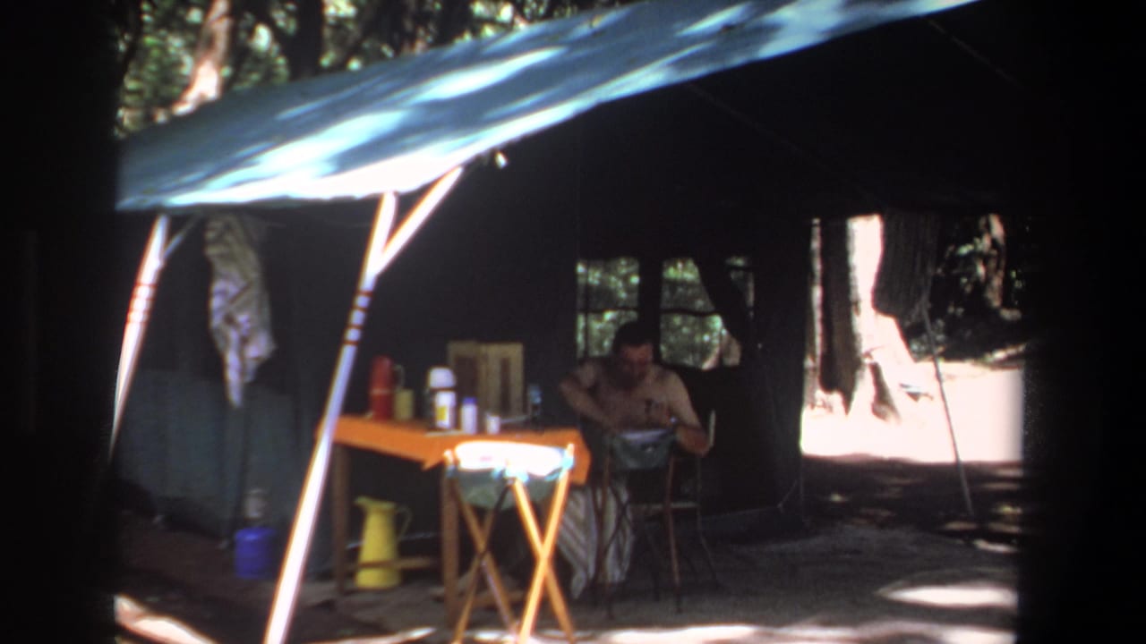Shaded campsite tent with a seated man and supplies on a small table.