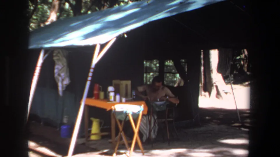 The final frame holds on the Pretoria forest tent, where the man sits near a small supply table under a blue tarp and dappled light.