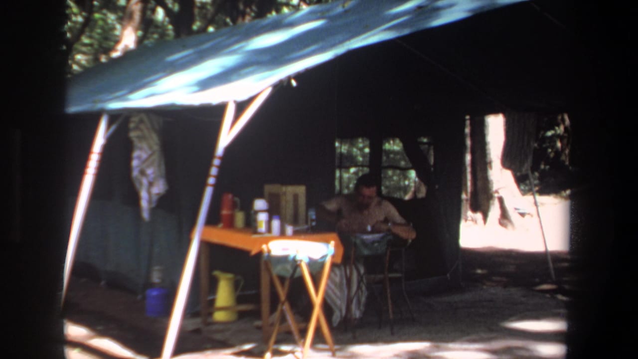 Man seated under a blue tarp shelter at a wooded campsite with a folding table and camping supplies.