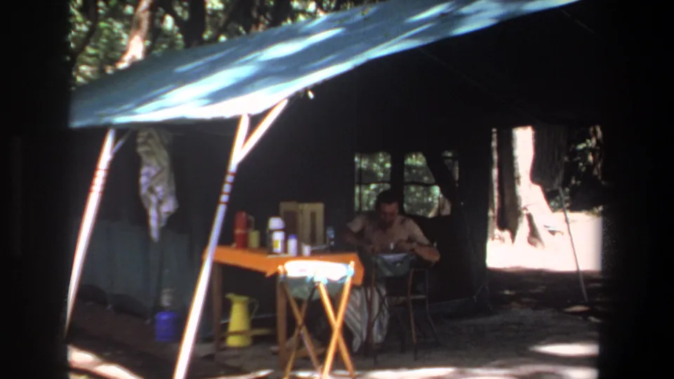 1969 Pretoria frame of a shaded campsite setup with blue tarp roofing, camp supplies on a folding table, and a seated man inside.