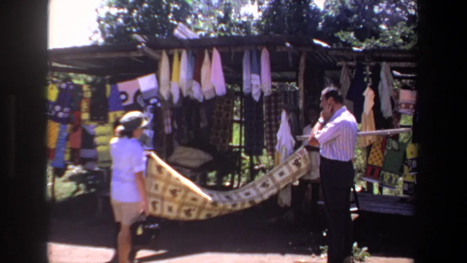 1969 Pretoria home-movie frame showing adults browsing textiles at an outdoor market stall with hanging fabrics and crafts.