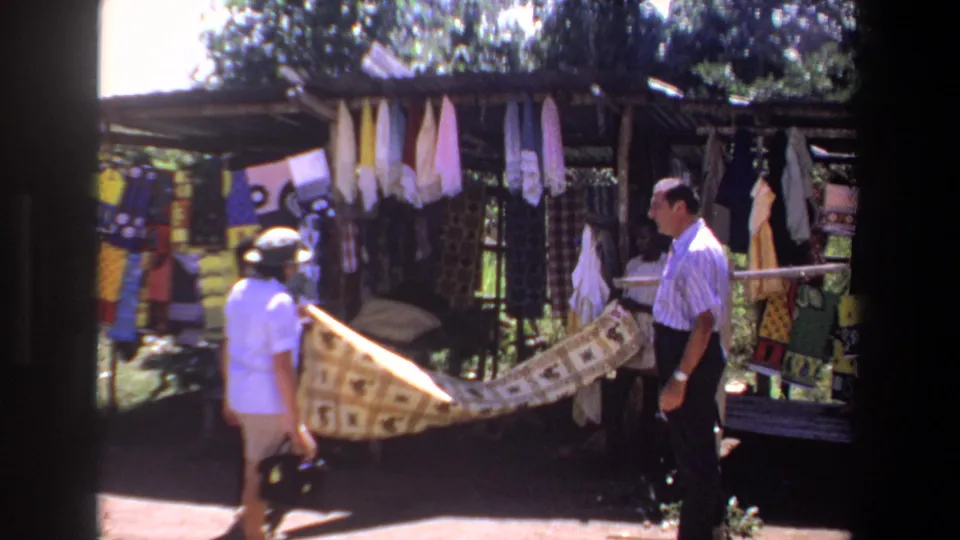 1969 Pretoria market frame with a man and another shopper beside a stall of hanging textiles and a patterned fabric in sunlight.