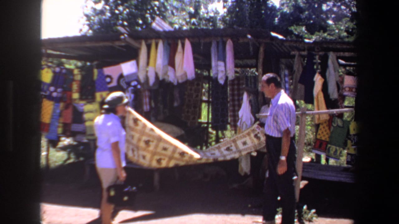 In 1969 Pretoria footage, a woman in a sun hat and a man stand by a shaded textile stall while a patterned fabric is held out between them.