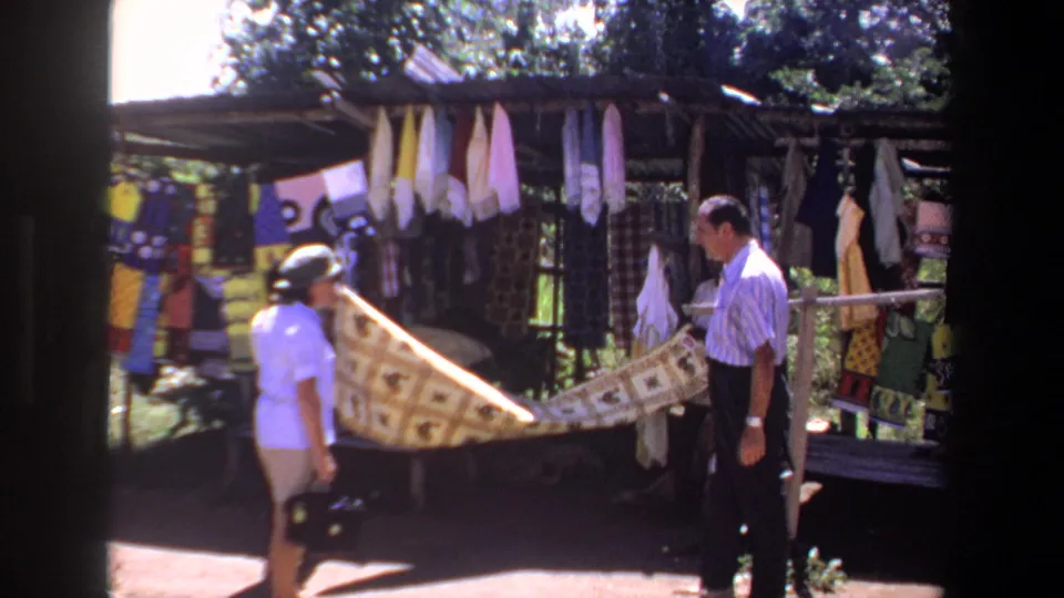 In 1969 Pretoria footage, a woman in a sun hat and a man stand by a shaded textile stall while a patterned fabric is held out between them.