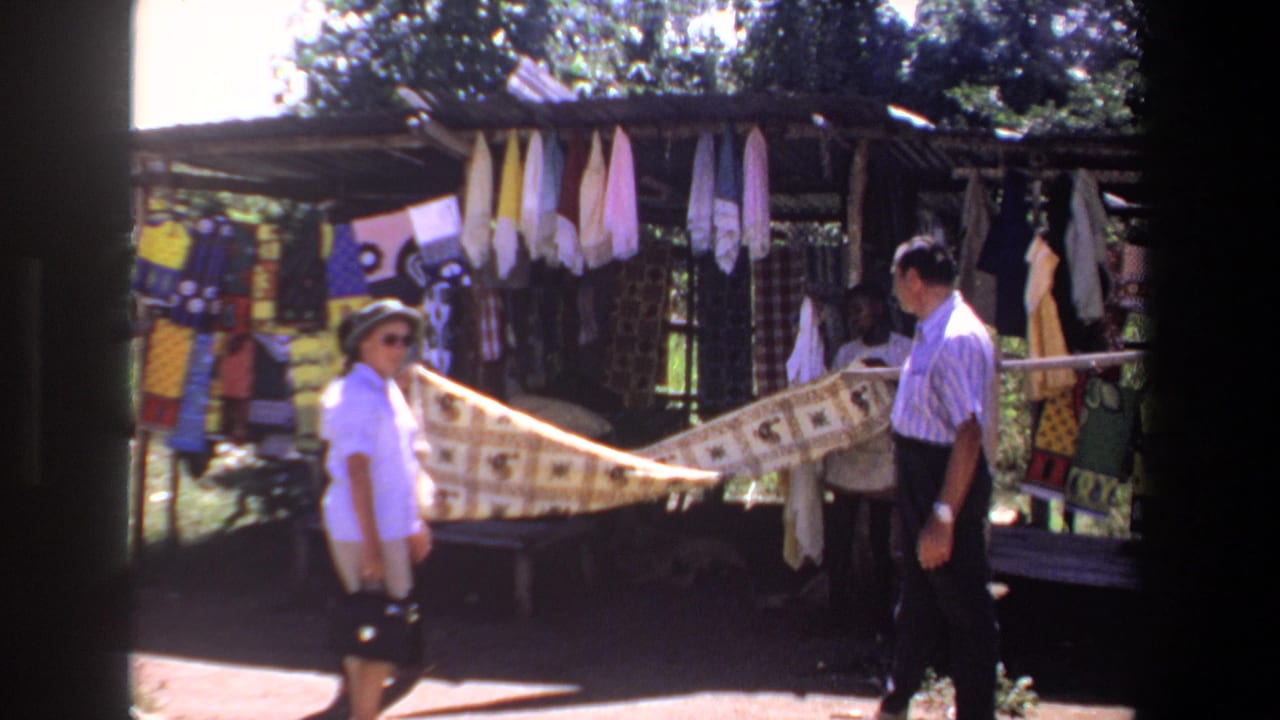The couple appears to inspect a long patterned textile at an outdoor stall in Pretoria, with hanging fabrics and greenery behind them.