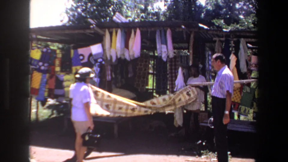 A vendor partly visible behind the cloth helps display a patterned fabric for two adult visitors at the Pretoria outdoor textile stall.