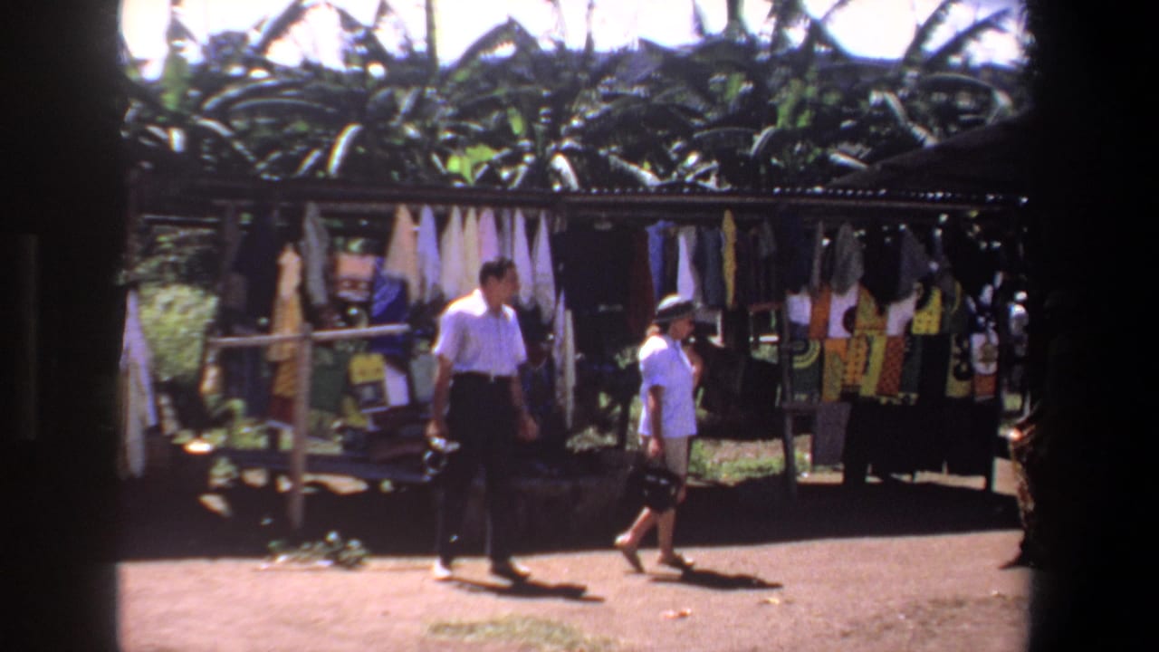 In supplied 1969 Pretoria footage, two tourists pass a sunlit outdoor stall displaying hanging textiles and garments.
