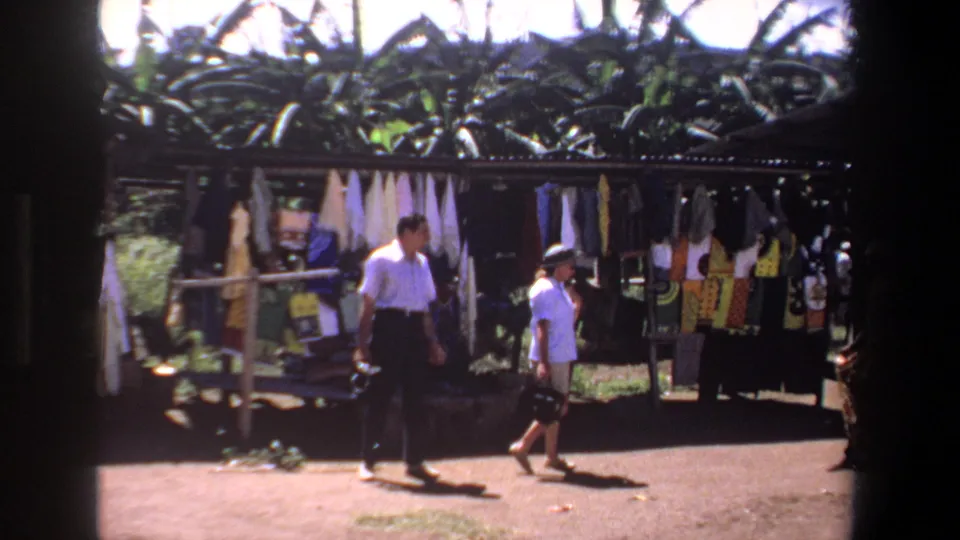 In supplied 1969 Pretoria footage, two tourists pass a sunlit outdoor stall displaying hanging textiles and garments.