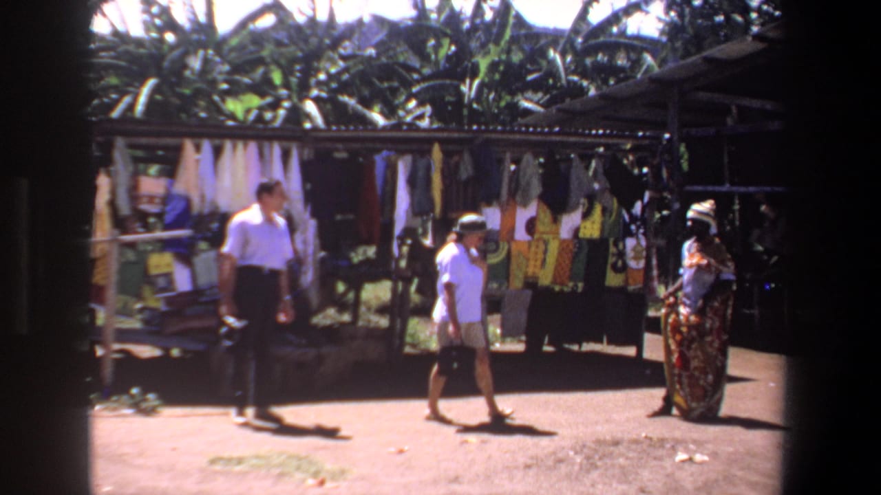 Two tourists approach a vendor near a stall with colorful textiles hanging in the background.