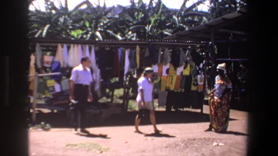 The visitors move toward a vendor beside a Pretoria market stall with garments and textiles hanging under shade.