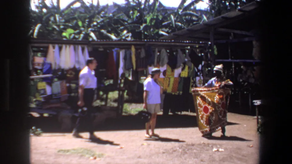 A vendor holds out a patterned textile for two tourists in the supplied 1969 Pretoria outdoor market scene.