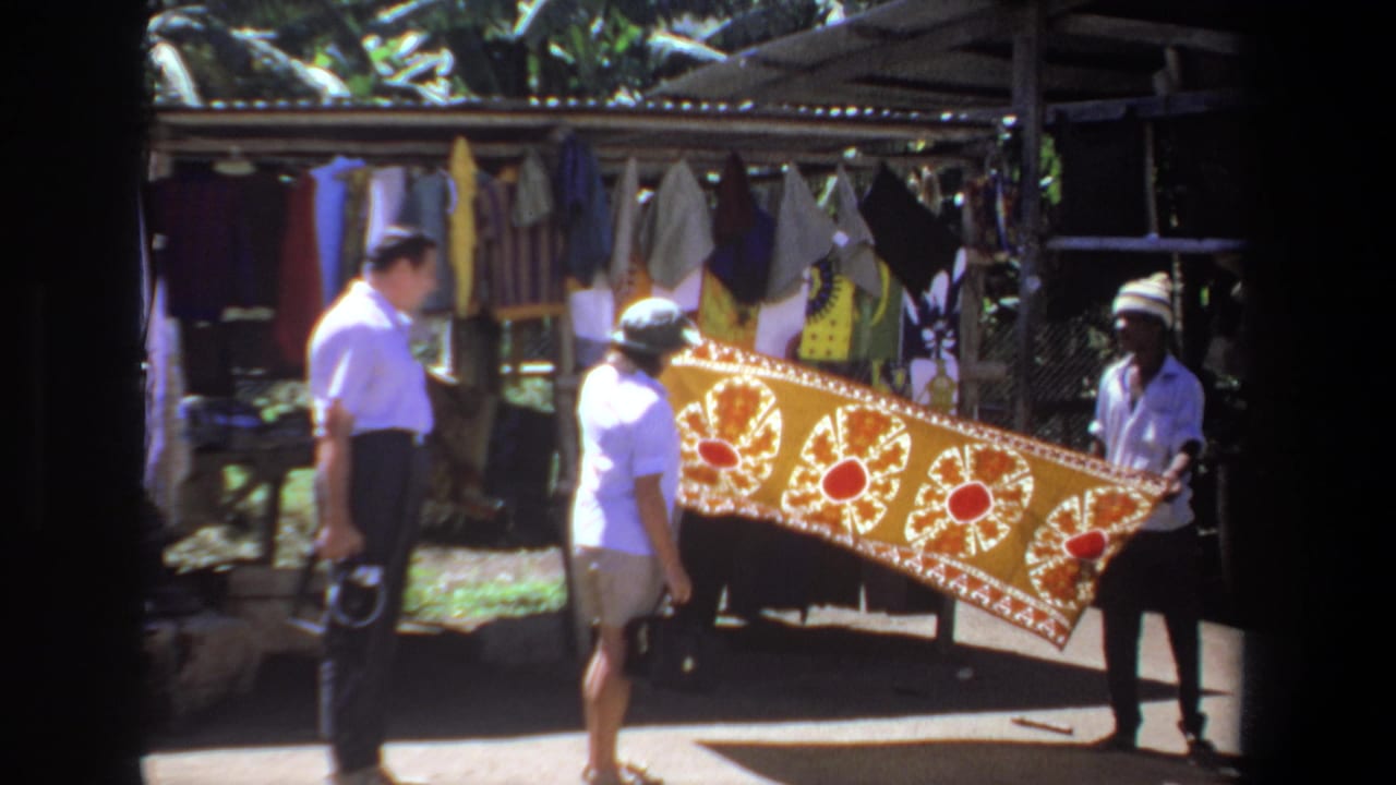 In Pretoria, South Africa, 1969, visitors look at a yellow textile with red and white patterns held at an outdoor market stall.