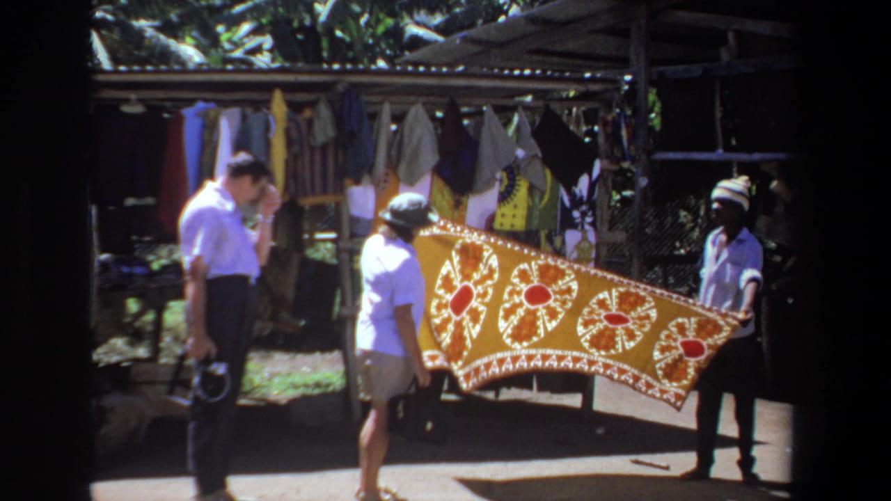 A vendor holds a yellow textile while two visitors stand beside the market stall.