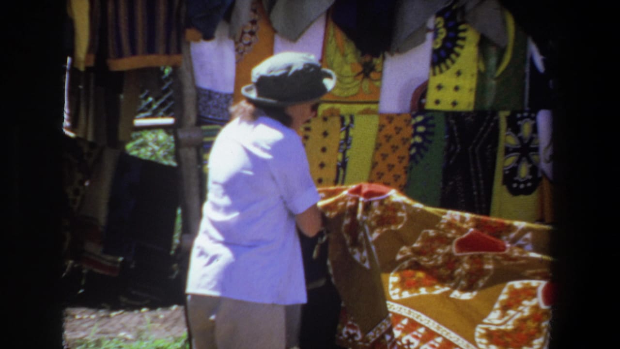 In Pretoria, South Africa in 1969, a woman in a gray hat and pale shirt looks over patterned fabric at an outdoor market stall.