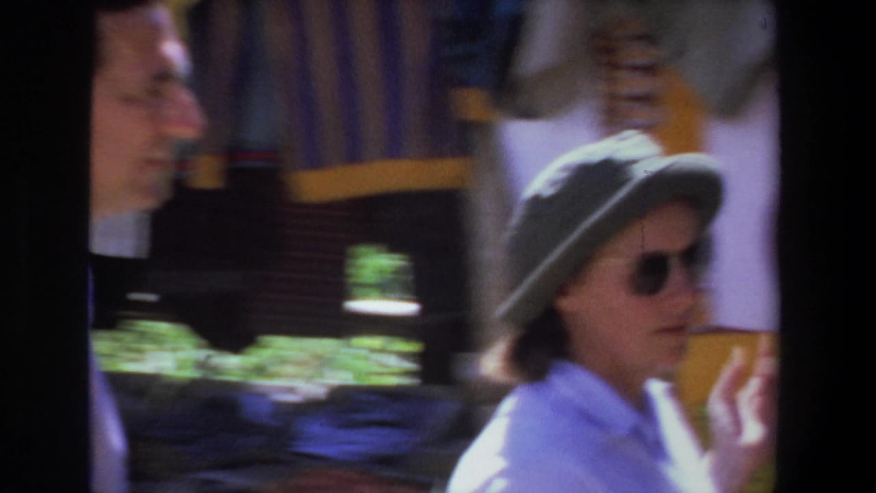 Woman in sunglasses and gray hat passes hanging textiles as a man moves behind her.